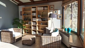 A cozy living room with wicker furniture, a bookshelf, and a view of the outdoors through a large window.