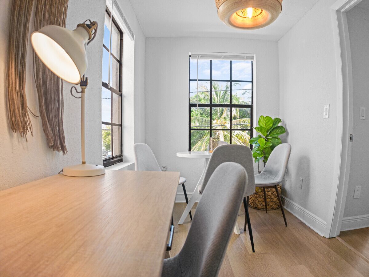 Un coin repas avec une table en bois, deux chaises, une lampe sur pied et une grande fenêtre donnant sur des palmiers.