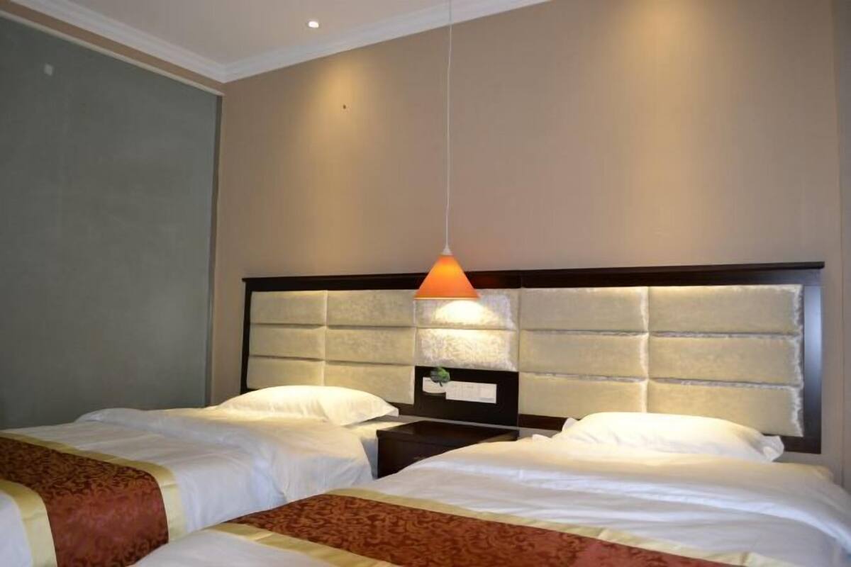 A hotel room with two beds, a headboard with a light fixture, and a nightstand with a lamp.