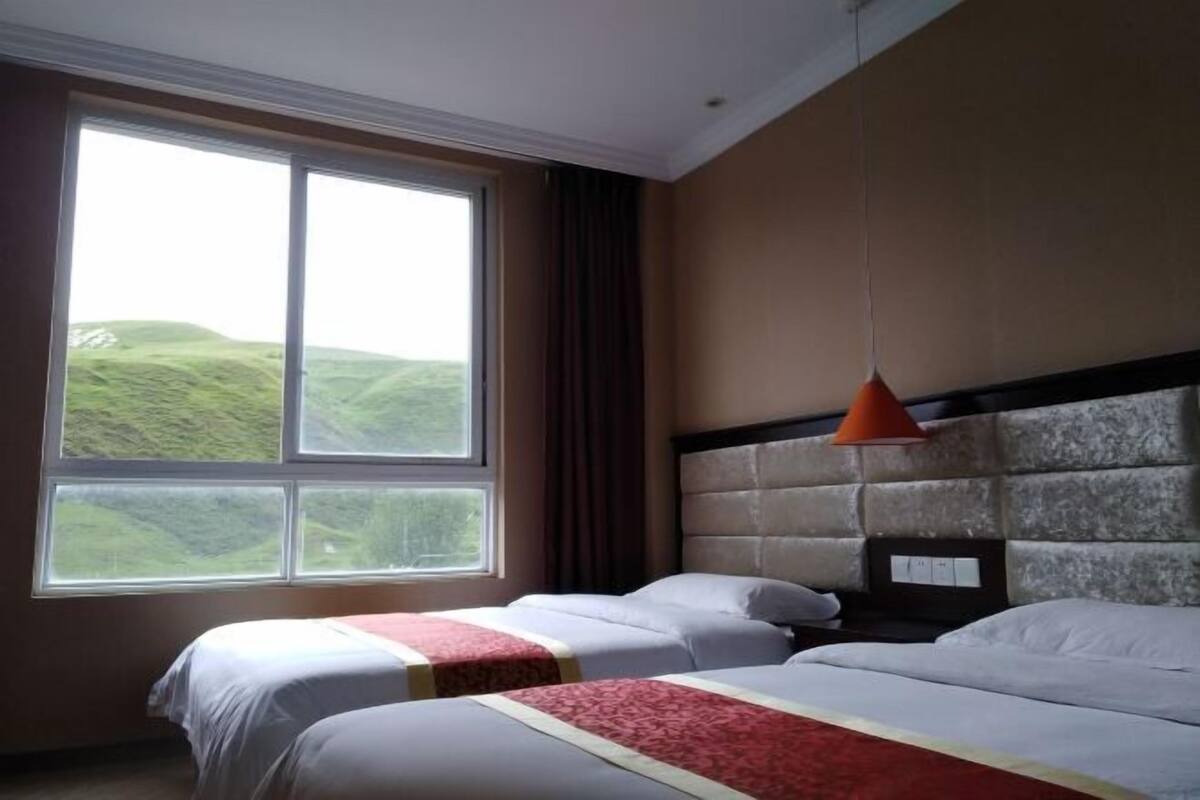 A hotel room with two beds, a large window with a view of green hills, a stone headboard, and a pendant light.