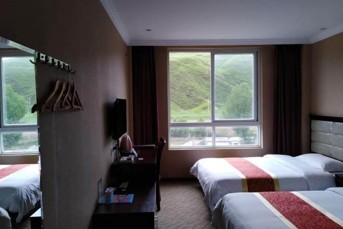 Une chambre d’hôtel avec deux lits, un bureau avec une chaise, une télévision et une grande fenêtre donnant sur des collines.