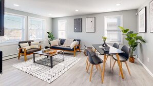 A modern living room with a sofa, coffee table, and dining table with chairs. There are large windows with blinds, a flat-screen TV, and framed artwork on the walls.