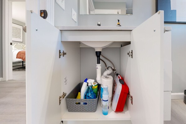 Ein Badezimmer mit einem weißen Waschbecken, einem Spiegel und verschiedenen Pflegeprodukten, darunter eine Handseifenflasche, eine Shampooflasche und ein roter Behälter.
