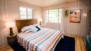 Un dormitorio con cabecera de madera, colcha estampada, dos lámparas de noche, una ventana con cortinas y una planta sujeta a la pared.