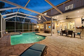 A covered outdoor swimming pool area with a patio, seating, and a view of the house.