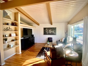A spacious living room with a wooden floor, a sofa, a coffee table, and a TV on a stand. There are shelves with decorative items and a world map on the wall.