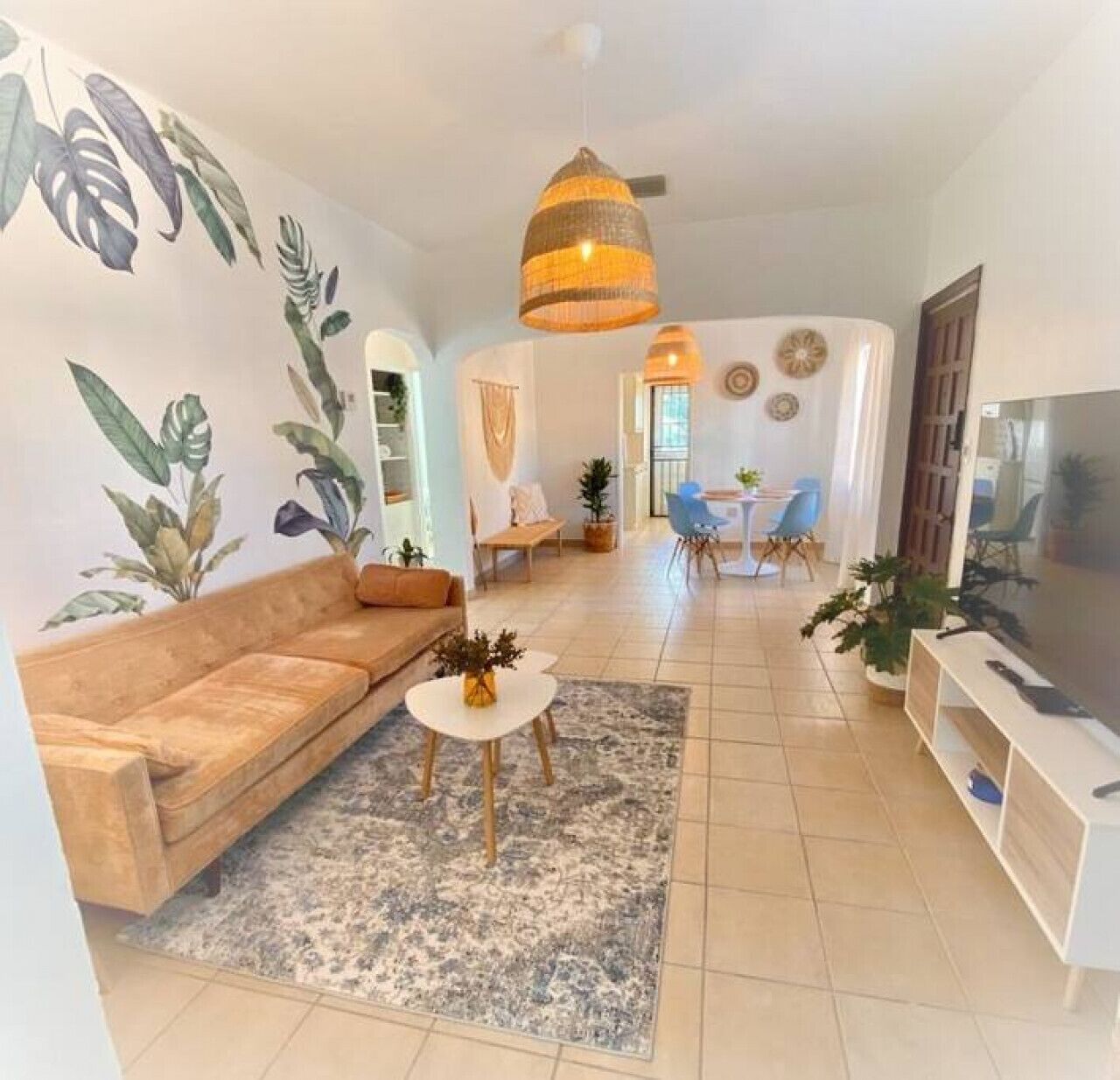 A modern living room with a sofa, coffee table, and TV stand. There are plants and a dining area in the background.