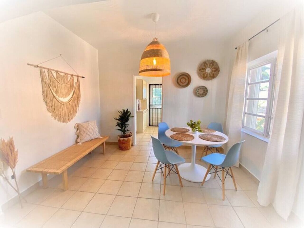 A bright, well-lit room with a round dining table, blue chairs, a hanging pendant light, a bench, and a potted plant.