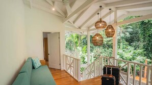 A room with a green sofa, wooden flooring, wicker pendant lights, and a view of lush greenery through a white balcony railing. - El Oasis Beachfront - Oceanfront Escape (Manzanillo)