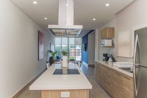 A modern kitchen with a central island, wooden cabinets, a stainless steel refrigerator, and a large window providing a view of the city.