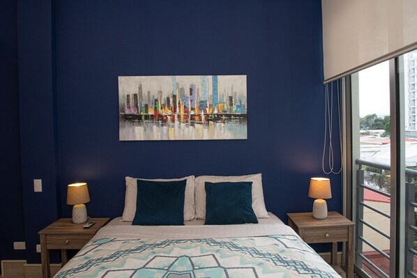 A bedroom with a cityscape painting on the wall, two bedside tables with lamps, and a patterned bedspread.