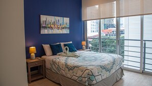 Ein Schlafzimmer mit einem großen Bett, zwei Nachttischen mit Lampen, einem Stadtbild an der Wand und Blick auf Gebäude durch das Fenster.