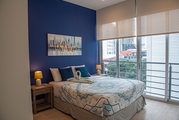A bedroom with a large bed, two bedside tables with lamps, a cityscape painting on the wall, and a view of buildings through the window.