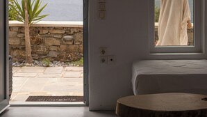 A room with a large window overlooking a stone wall and a palm tree, a bed with a white headboard, and a round wooden table. - Villa 9 - Sea View, Private Jacuzzi for 5, Shared Pool,Gym,Bar (Mykonos)