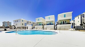 A swimming pool area with a clear blue sky, surrounded by modern buildings with balconies and a flat roof.
