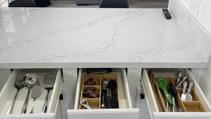 Een keuken met een wit marmeren aanrecht, lades met keukengerei, en een grijze bank op de achtergrond.