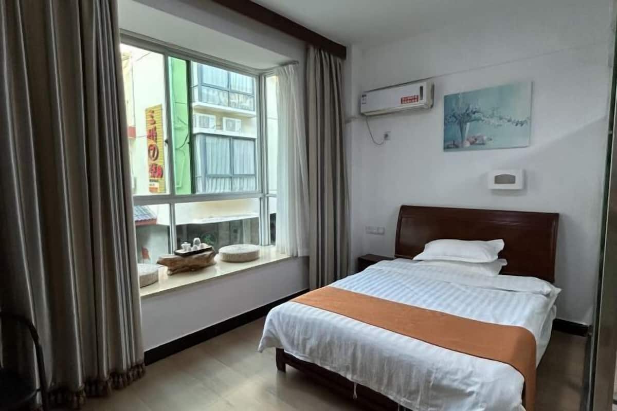 Una habitación de hotel con cama, ventana con cortinas, aire acondicionado y una obra de arte colgada en la pared.