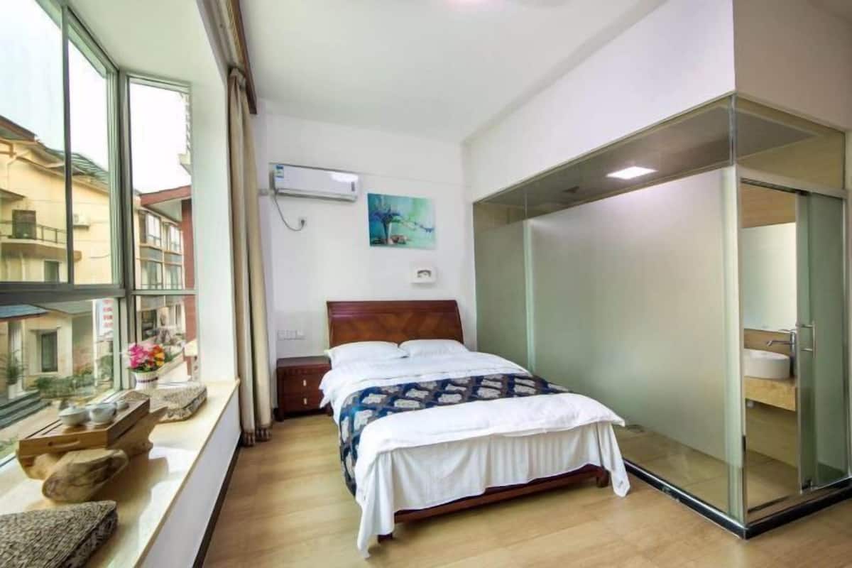 Un dormitorio con cama, ventana con vistas a edificios, mesita de noche con lámpara y puerta corredera de vidrio que da a un baño.