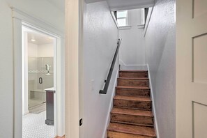 Eine enge Treppe mit hölzernen Stufen und einer Handstange, die zu einem Raum mit Duschkabine aus Glas und weiß gefliestem Boden führt.