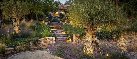 A garden pathway leading to a house with stone steps and a stone wall, surrounded by various plants and flowers.