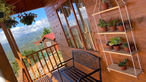Uma varanda de madeira com um banco de metal, plantas em vasos e vista para a paisagem ao redor.