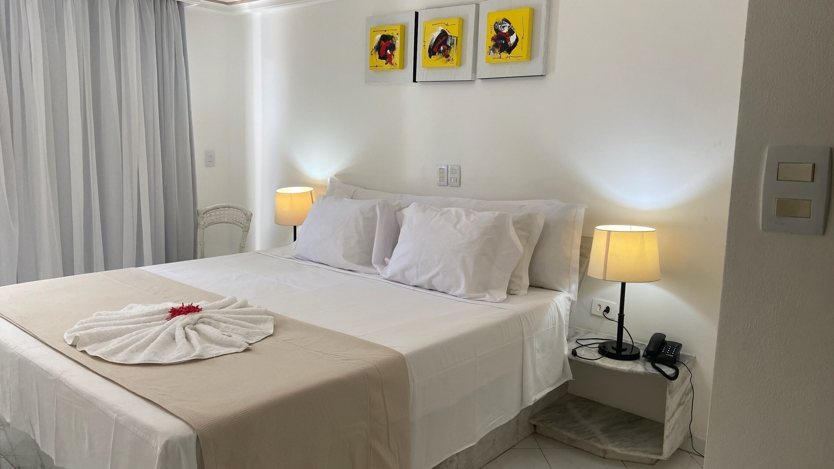 Habitación de hotel con una cama bien hecha, dos lámparas de noche, un teléfono y tres obras de arte enmarcadas en la pared.