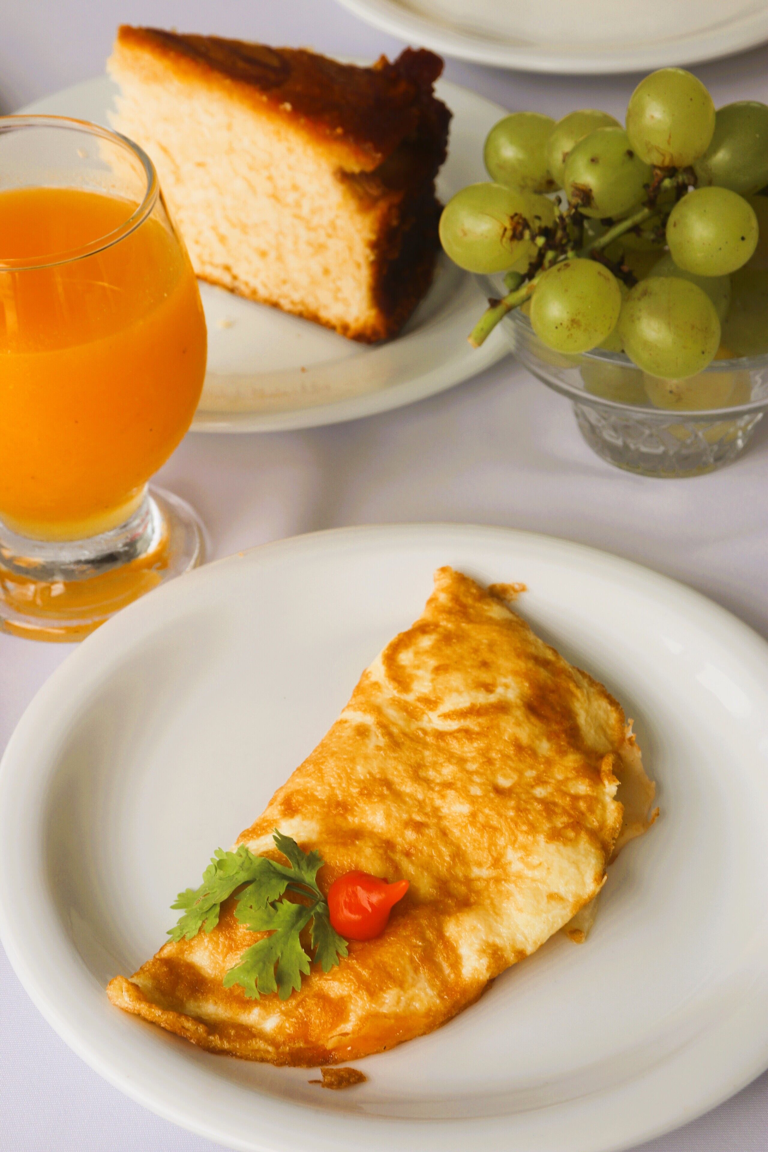 Un plato con una tortilla, una porción de pastel, un vaso de jugo de naranja y un bol de uvas.