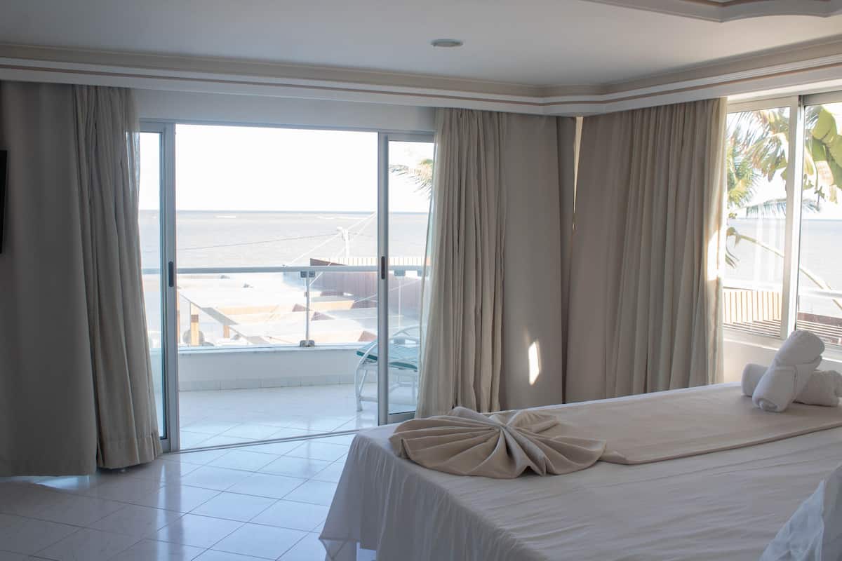 A hotel room with a large bed, a view of the beach, and a balcony with a glass door.