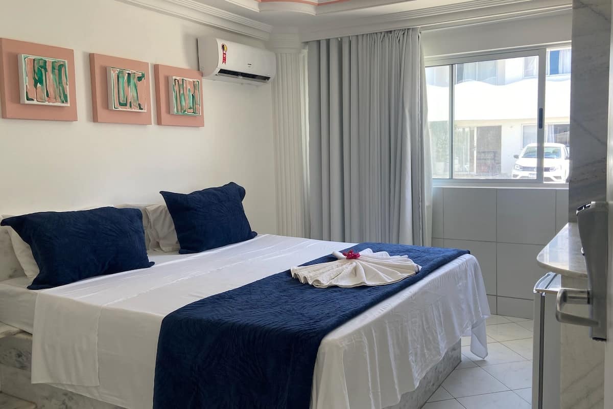 A hotel room with a bed, pillows, a towel, an air conditioner, a window with curtains, and artwork on the wall.