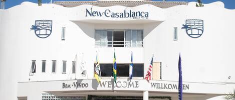The exterior of New Casablanca Praia Hotel with a welcoming sign, flags, and a clear blue sky.