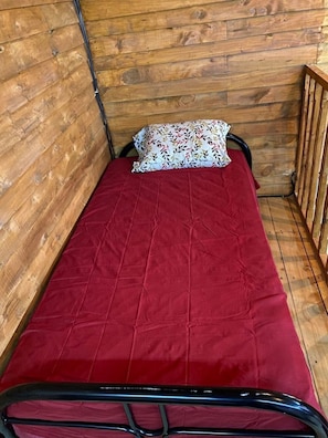 A small, single-bed room with a wooden wall and floor, a red bedspread, and a patterned pillow.