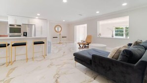 A modern living room with a dark sofa, a round coffee table, and a kitchen area with white cabinets and a marble floor. - The Villa #1 (North Palm Beach)