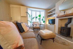A living room with a sofa, armchair, ottoman, and a television. There is a fireplace and a window with blinds. - Juniper Cottage is a quaint, two-bedroom Grade II listed property featuring white wooden cladding. (Whitstable)