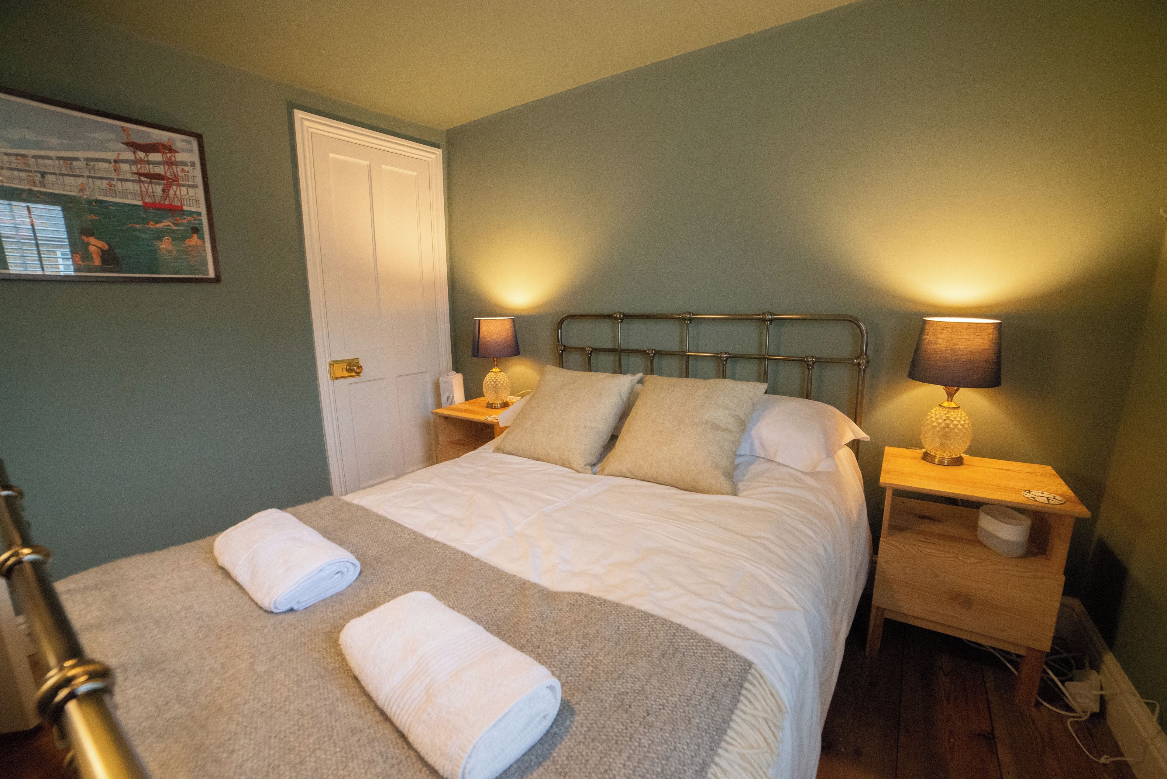 A neatly made bed with two pillows and two rolled towels, a nightstand with a lamp, and a door to the left.