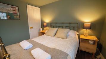 A neatly made bed with two pillows and two rolled towels, a nightstand with a lamp, and a door to the left.
