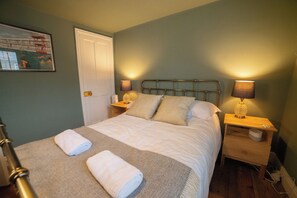 A neatly made bed with two pillows and two rolled towels, a nightstand with a lamp, and a door to the left. - Juniper Cottage is a quaint, two-bedroom Grade II listed property featuring white wooden cladding. (Whitstable)