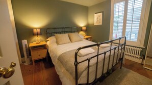 Un dormitorio con estructura de cama de metal, dos mesitas de noche con lámparas, un radiador y una ventana con persianas.