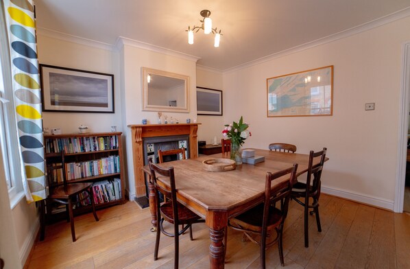 A dining room with a wooden table, chairs, a bookshelf, a fireplace, and framed artwork on the walls.