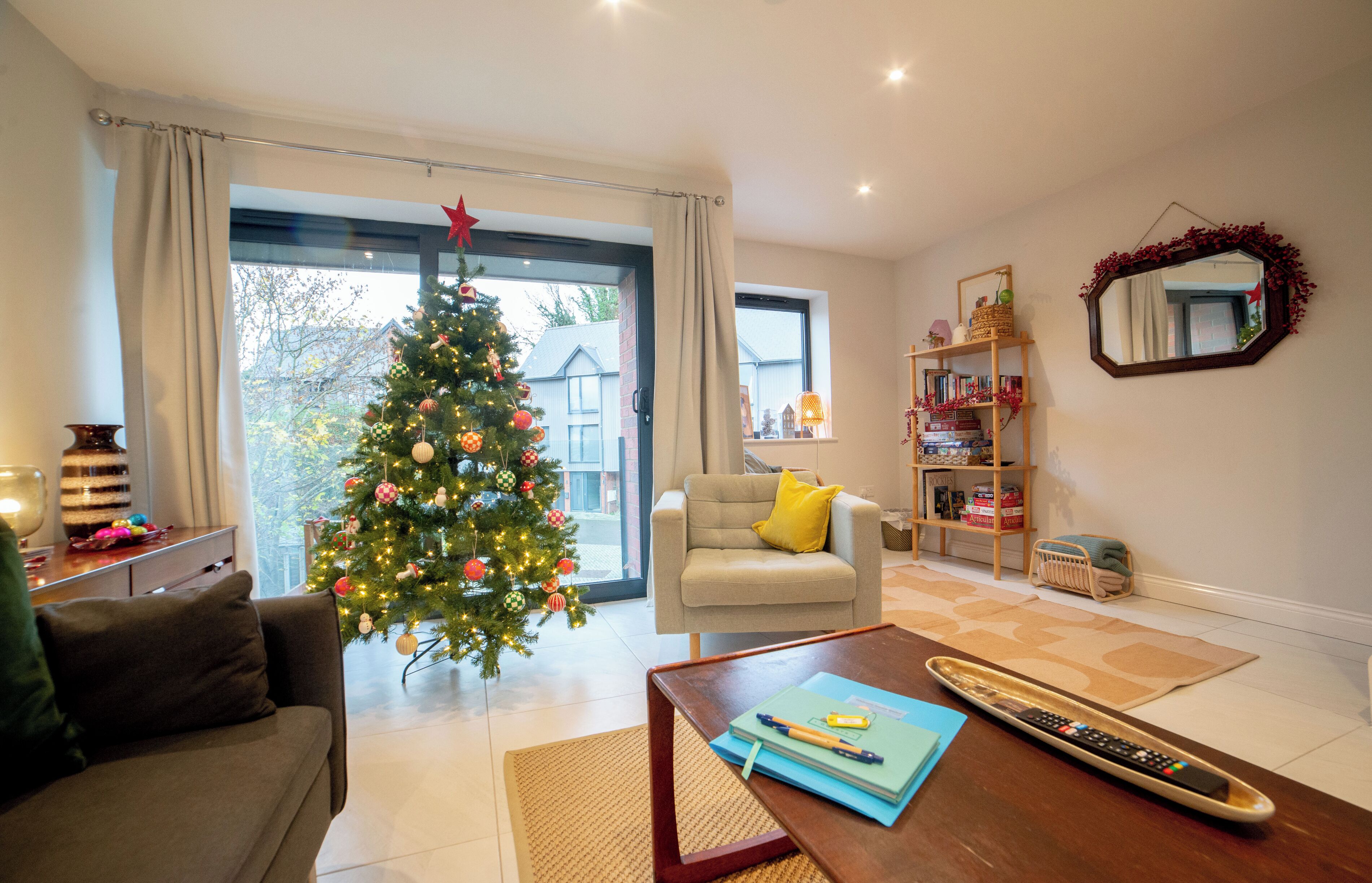 A living room with a Christmas tree, a sofa, a coffee table, a bookshelf, and a window with curtains.