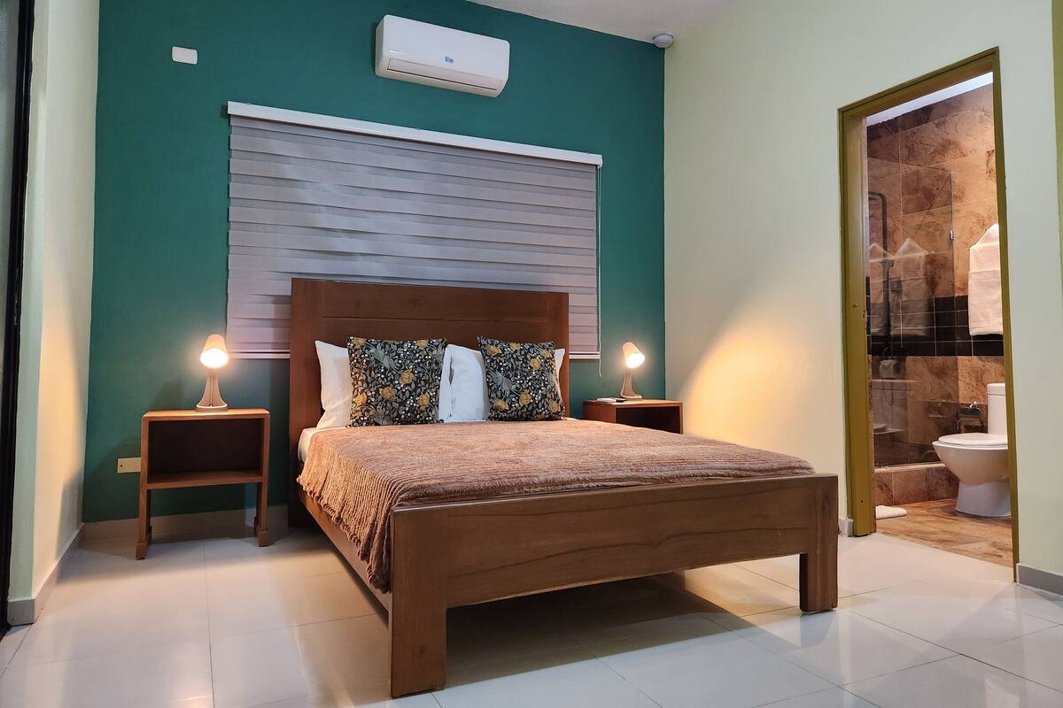 A bedroom with a wooden bed, two bedside lamps, a wall-mounted air conditioner, and a bathroom visible through an open door.