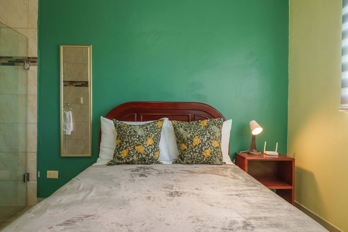 A bedroom with a bed, two pillows, a nightstand with a lamp, a mirror, and a shower area.