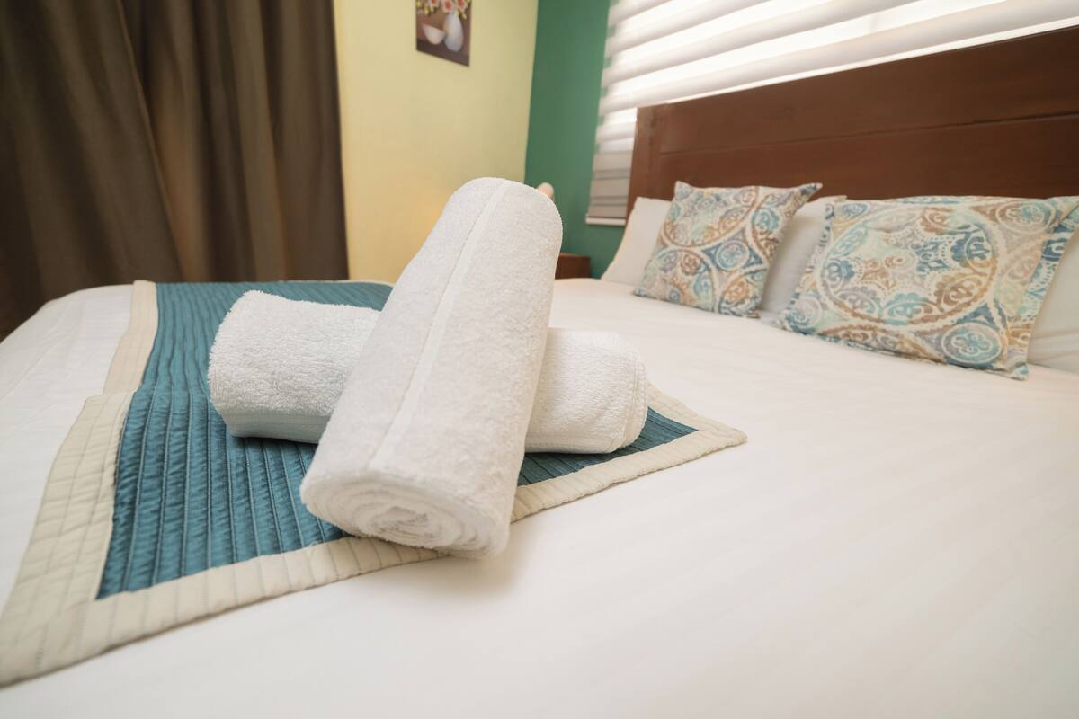 A neatly made bed with a rolled white towel, a teal bedspread, and patterned pillows.