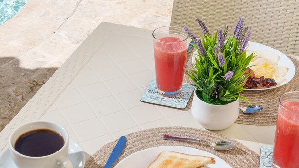 A dining table with breakfast items, a coffee cup, and a drink by a poolside. - La Casona MC (La Romana)