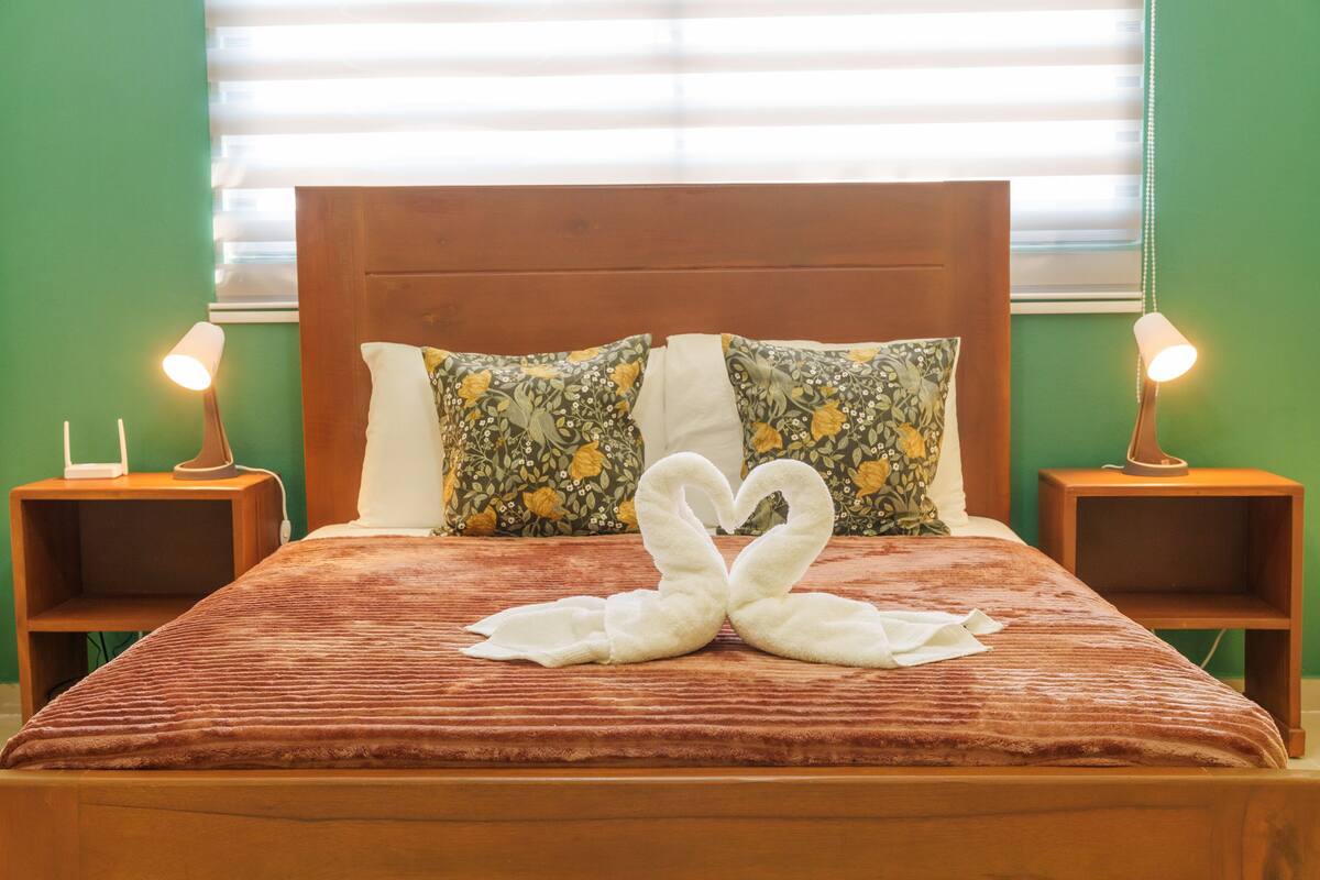 A neatly made bed with a swan-shaped towel arrangement, green walls, and bedside lamps.