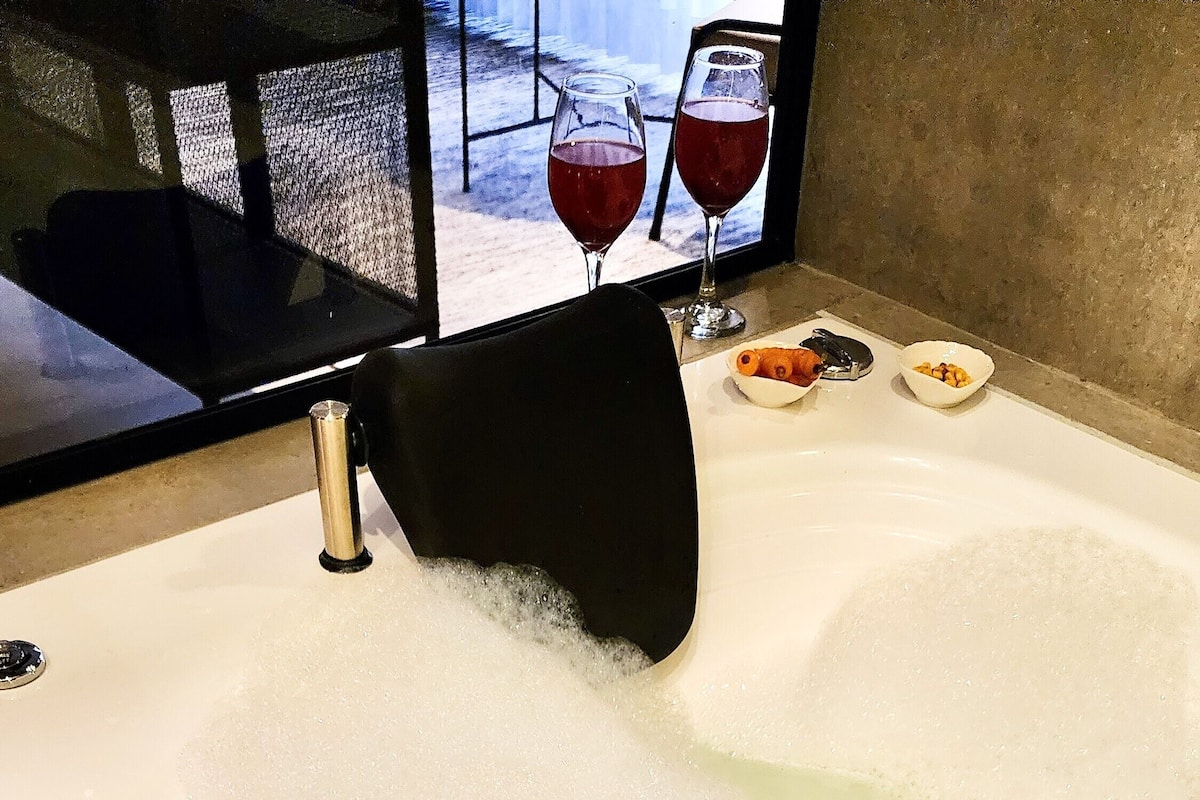 A bathtub with running water, a black chair, and two glasses of red wine on a table.