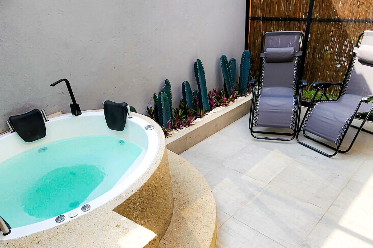 A hot tub with a black faucet, surrounded by a stone border. There are two lounge chairs with cushions.