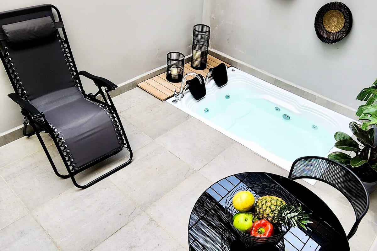 A pool area with a black lounge chair, a hot tub, and a table with fruits.