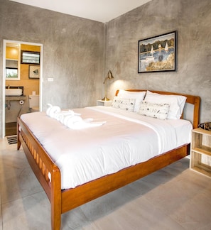 Un dormitorio con una cama de madera, una mesita de noche, una lámpara, un cuadro en la pared y un baño visible a través de una puerta abierta.