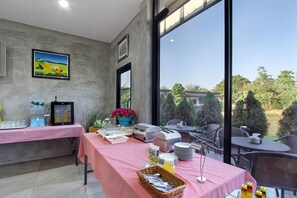 A dining area with a table set for breakfast, a large window, and a painting on the wall. - White Knot Khao Yai (Pak Chong)
