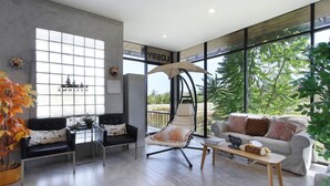 A modern living room with a swing chair, two sofas, a coffee table, and large windows offering a view of greenery. - White Knot Khao Yai (Pak Chong)